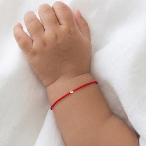 Op de afbeelding: Een dunne rode koordarmband met een enkele kleine, heldere, rondgeslepen edelsteen in een metalen zetting. De armband wordt om een pols gedragen en toont een minimalistisch ontwerp met een felrood koord en een centraal steentje.