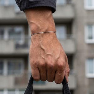 May include: A minimalist bracelet with a thin, grey cord and a small, round, silver charm. The bracelet is worn on a wrist. The background shows a blurred building.