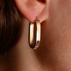 May include: Gold hoop earrings with a rectangular shape. The earrings are worn in a person's ear.