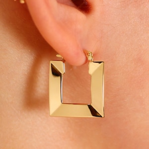 May include: Gold square hoop earrings with a slightly angled edge. The earrings are worn in a person's ear.