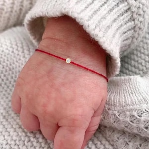 Pode incluir: Uma delicada pulseira de fio vermelho com uma pequena pedra preciosa redonda e transparente, engastada numa base dourada. O fio fino envolve o pulso, destacando o design minimalista da pedra central em contraste com o fio vermelho vivo.