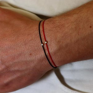 Może przedstawiać: Dwie minimalistyczne bransoletki, jedna czarna i jedna czerwona, noszone na nadgarstku. Każda bransoletka ma mały, błyszczący kamień szlachetny. Bransoletki są proste i eleganckie, odpowiednie do codziennego użytku.