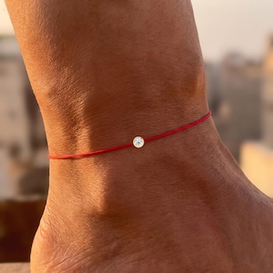 May include: A thin red cord anklet featuring a single round clear gemstone set in a gold-toned bezel. The delicate string wraps around the ankle, holding the small, circular stone centred as a minimalist focal point against the skin.