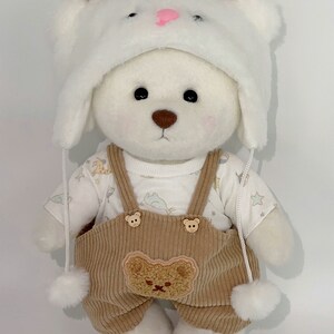 May include: White plush teddy bear wearing a white furry hat with brown ears and a brown corduroy overall with a bear patch. The teddy bear is wearing a white t-shirt with a star pattern.