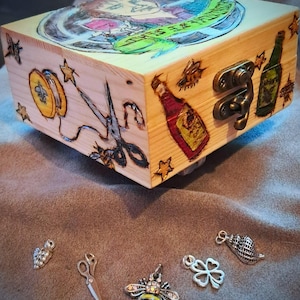 May include: A wooden box with hand-drawn illustrations and metal clasp. The box features images of scissors, a bottle, and a cartoon figure. Several small charms, including a bee, scissors, and a four-leaf clover, are scattered below.
