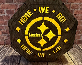 Könnte beinhalten: Ein Schild aus Holz mit einem gelben Steelers-Logo und dem Text "Here We Go!" um das Logo herum. Das Schild ist von hinten beleuchtet, wodurch das Logo und der Text leuchten.