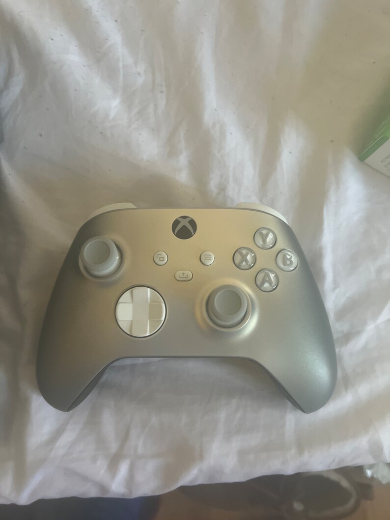 May include: Silver Xbox controller with white accents. The controller features the Xbox logo, a directional pad, and various buttons. The design is sleek and modern, with a metallic finish.