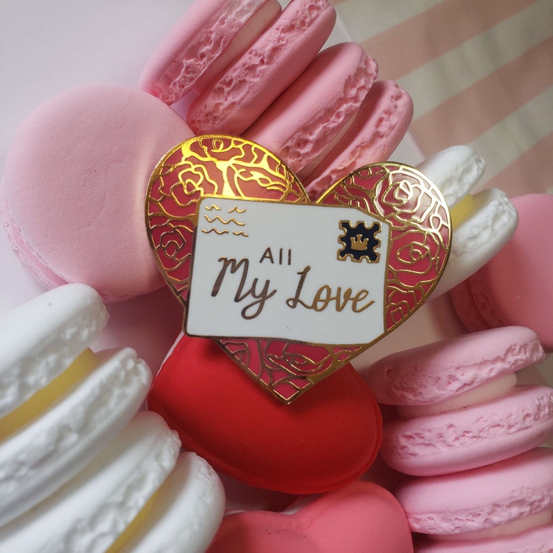 以下が含まれることがあります： 白い封筒のデザインと「All My Love」の文字が書かれた、金色のハート型のピン。ピンはピンクと白のマカロンで囲まれています。