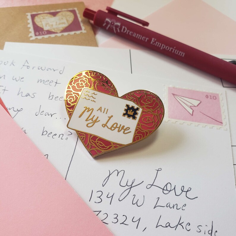 以下が含まれることがあります： 白い背景に「All My Love」と書かれた、ハート型の金と赤のエナメルピン。ピンは、「My Love 134 W Lane 72324, Lake side」と黒インクで書かれた白いポストカードの上に置かれています。