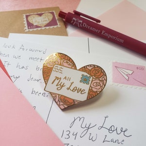 以下が含まれることがあります： 白い封筒のデザインと「All My Love」の文字が書かれた、金色のハート型のエナメルピン。封筒には、王冠とピンクのバラのデザインが描かれた青い切手が貼られています。ピンは、手書きのメモが書かれた白い背景の上に置かれています。