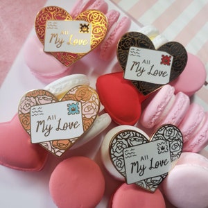 Puede incluir: Cuatro pines de esmalte en forma de corazón con detalles dorados. Cada pin presenta un sobre blanco con el texto "All My Love" y un sello decorativo. Los pines están dispuestos sobre un fondo de macarons rosas y blancos.
