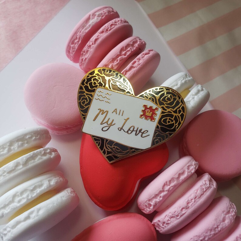 以下が含まれることがあります： 黒い花柄の金色のハート型のピンと、「All My Love」と書かれた白い封筒。ピンは赤いハート型の物体のの上に置かれています。背景にはピンクと白のマカロンがあります。