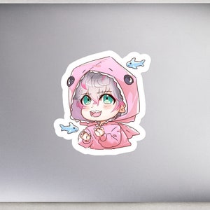 May include: A sticker featuring a cartoon character wearing a pink shark costume with a white and grey face. The character has green eyes and is smiling. There are small blue fish around the character.