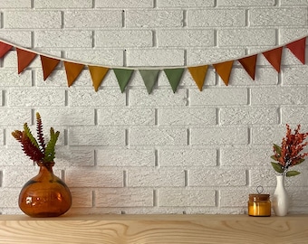 Op de afbeelding: Een witte bakstenen muur met een herfstbanner van oranje, rode en groene stoffen driehoeken. Twee vazen met herfstbladeren staan op een houten plank onder de banner. Een vaas is van amberkleurig glas en de andere van wit keramiek.