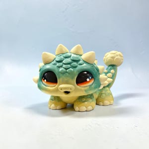 Puede incluir: Figura de dinosaurio pintada a mano en tonos verde azulado y amarillo. La figura presenta grandes ojos expresivos y un diseño detallado con púas y una maza en la cola. Un objeto decorativo encantador, ideal para exhibir. Mide unos 10 cm de largo.