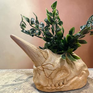 May include: A beige, textured planter shaped like a fossilized dinosaur skull with a prominent horn and visible cracks. The skull features a row of small, pointed teeth along the jawline and contains a cluster of vibrant green artificial leaves.