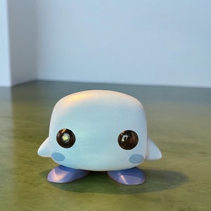 May include: A small, white, ceramic figurine with a rounded shape and large, black eyes. The figurine has a light blue and purple paint detail on the bottom.