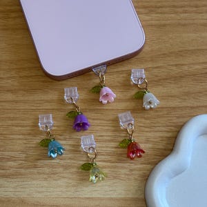 Può includere: Una collezione di ciondoli per telefono a forma di fiore colorati. Ogni ciondolo presenta un piccolo fiore traslucido nelle tonalità del rosa, viola, blu, giallo, rosso e bianco, con una foglia verde e dettagli in metallo dorato. I ciondoli sono attaccati a connettori di plastica trasparente.