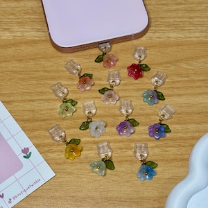 Può includere: Una collezione di ciondoli per telefono a forma di fiore colorati. Ogni ciondolo presenta un piccolo fiore traslucido nei toni del rosa, rosso, giallo, blu e viola, con accenti di foglie verdi. I ciondoli sono collegati a una spina trasparente.