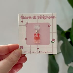 Puede incluir: Un charm de teléfono de oso de goma rosa y rojo. El charm está sujeto a un lazo de plástico transparente. La tarjeta de respaldo tiene el texto "Charm de téléphone" y nombres de usuario de redes sociales. El fondo es un patrón de cuadrícula con corazones y flores.