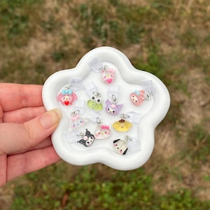 Puede incluir: Una bandeja blanca en forma de flor que contiene varios pequeños amuletos de colores. Los amuletos presentan varios personajes y diseños de dibujos animados, incluyendo gatos, lazos y corazones. Cada amuleto está unido a un clip de plástico transparente.