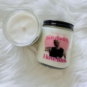 May include: Two glass jar candles with white wax. One candle is open, showing the wax surface. The other candle has a black lid and a pink label with the text "but daddy, i love him" and a photo of a man.