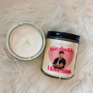 Puede incluir: Dos velas en frascos de vidrio con cera blanca. Una vela está abierta, mostrando la mecha. La otra vela tiene una tapa negra y una etiqueta con el texto "but daddy, i love him" sobre un corazón rosa y una foto de un hombre.