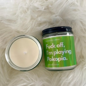 May include: Two glass jar candles with white wax and a wick. One candle is open, the other has a black lid and a green label with the text "Fuck off, I'm playing Pokopia."
