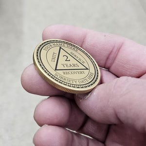 May include: A gold-colored sobriety coin with the text "YOUR NAME HERE" around the edge. The center features a triangle with "2 YEARS" and the words "UNITY", "SERVICE", and "RECOVERY". The coin also has the text "SOBRIETY DATE HERE".