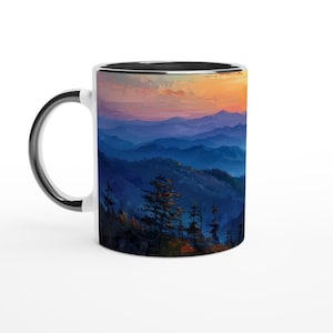 May include: White ceramic mug with a black rim, featuring a scenic image of a mountain range at sunset. The mountains are layered in shades of blue and purple, with a few trees silhouetted in the foreground.