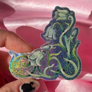 May include: A die-cut sticker featuring a watercolor illustration of bluebell flowers with green stems and leaves. The sticker has a holographic finish that creates a shimmering, multi-colored effect across the dark blue background.