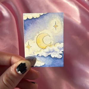 Pode incluir: Uma pequena pintura em aquarela de uma cena do céu noturno. A obra apresenta uma lua crescente amarela, duas estrelas amarelas e nuvens brancas e fofas contra um fundo azul e roxo. A pintura é segurada por uma pessoa.