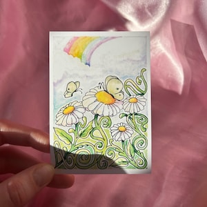 Könnte beinhalten: Ein Aquarellgemälde mit Gänseblümchen mit gelben Zentren und weißen Blütenblättern. Zwei Schmetterlinge flattern über den Blumen, und ein Regenbogen spannt sich über den Himmel. Das Kunstwerk befindet sich auf einer rechteckigen Karte.