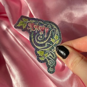 May include: A decorative sticker featuring a floral design with pink heart-shaped flowers, green leaves, and swirling vines. The sticker has a dark blue background with glitter accents and a glossy finish.