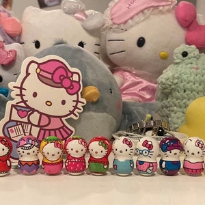 Könnte beinhalten: Ein Set mit 12 Hello Kitty Nistpuppen, jede mit einem anderen Design und einer anderen Farbe. Die Puppen sind in einer Reihe auf einer weißen Oberfläche angeordnet. Die Puppen sind alle etwa 5 cm groß.
