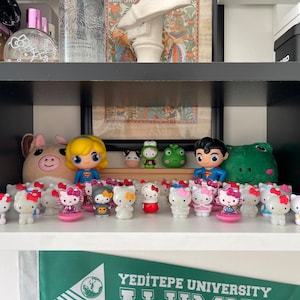 Könnte beinhalten: Ein Regal zeigt eine Sammlung von Figuren, darunter Hello Kitty-Charaktere mit roten Schleifen, Superheldenfiguren und ein Plüschschwein. Das Regal enthält auch Parfümflaschen, ein gerahmtes Bild und eine Casio-Uhrenbox.