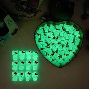 Könnte beinhalten: Eine herzförmige Schale gefüllt mit im Dunkeln leuchtenden Hello Kitty-Anhängern. Auf einer weißen Oberfläche sind außerdem 16 einzelne Anhänger in einem Raster angeordnet.