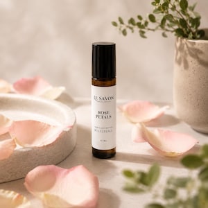 Op de afbeelding: Een bruine glazen rollerballfles met een zwarte dop, met het opschrift "Rose Petals" en "100% Handcrafted Rollerball". De fles is omgeven door roze rozenblaadjes en staat op een lichtgekleurd oppervlak. Het product is van "Le Savon Candles and Soaps".