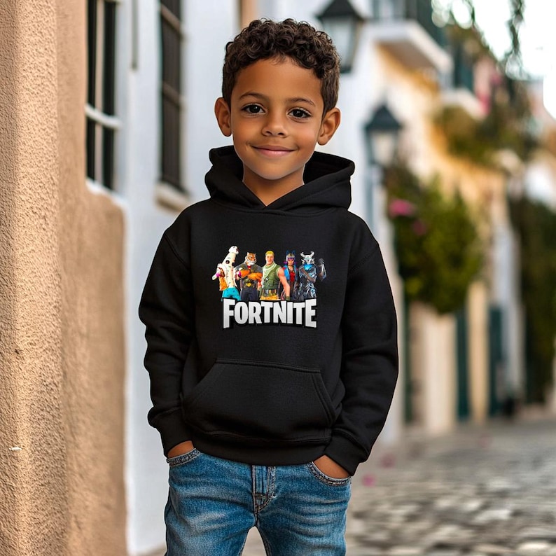 May include: Black hooded sweatshirt with a Fortnite character graphic and the word "FORTNITE" in white. The sweatshirt has a front pocket and is worn by a young person wearing blue jeans. The background is a blurred street scene.