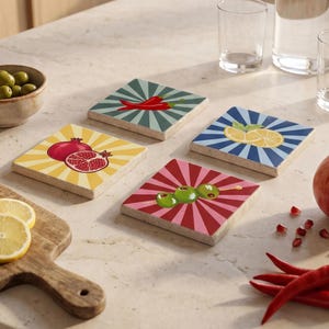 May include: Set of four square coasters with colorful fruit and vegetable designs. Each coaster features a different graphic: a pomegranate, lemons, olives, and a chili pepper. The coasters have a retro sunburst pattern in the background.