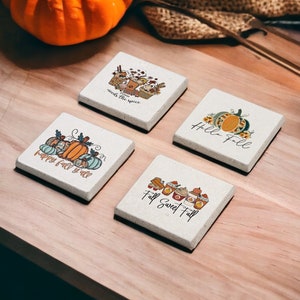 Könnte beinhalten: Vier quadratische Untersetzer mit Herbstmotiven. Die Untersetzer sind weiß mit braunen Rändern und zeigen Illustrationen von Kürbissen, Kaffeetassen und die Worte "Happy Fall Y'all", "Hello Fall", "Fall Sweet Fall" und "Sweeter the Spice".