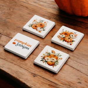 Pode incluir: Conjunto de quatro  tapetes de cerâmica branca com designs de outono. Um tapete apresenta o texto "pumpkin season" com um pequeno ícone de abóbora. Os outros três tapetes apresentam um buquê de flores laranja e brancas com abóboras.