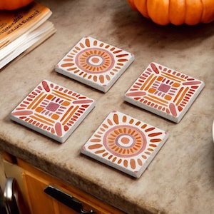 May include: Four square ceramic coasters with a white background and orange and pink geometric designs. The coasters are on a brown countertop.