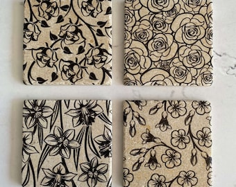 Op de afbeelding: Set van vier vierkante onderzetters met zwarte bloemenmotieven op een beige achtergrond. De ontwerpen omvatten rozen, narcissen en andere bloemenmotieven. De onderzetters zijn gemaakt van natuursteen en hebben een rustieke, landelijke esthetiek.