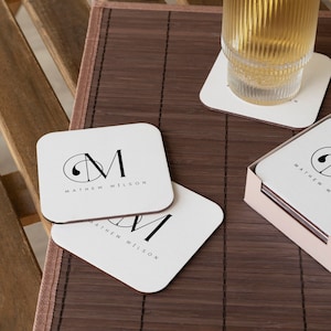 May include: Set of four white square coasters with black text and a monogram design. The coasters feature the name "MATTHEW WILSON" and a stylized "M" with a circle around it. The coasters are on a brown bamboo mat.