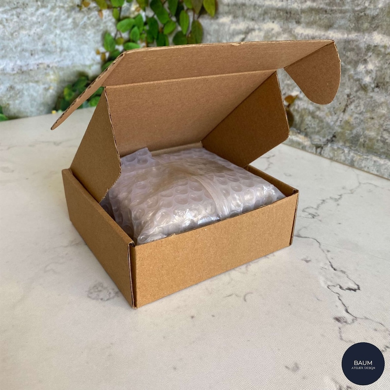 K&ouml;nnte beinhalten: Ein brauner Karton mit offenem Deckel, der ein wei&szlig;es Luftpolsterverpackungspaket im Inneren zeigt. Der Karton steht auf einer wei&szlig;en Oberfl&auml;che.