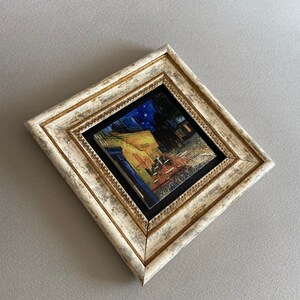 May include: A framed print of a painting depicting a cafe at night with people sitting at tables outside. The frame is white with gold accents.