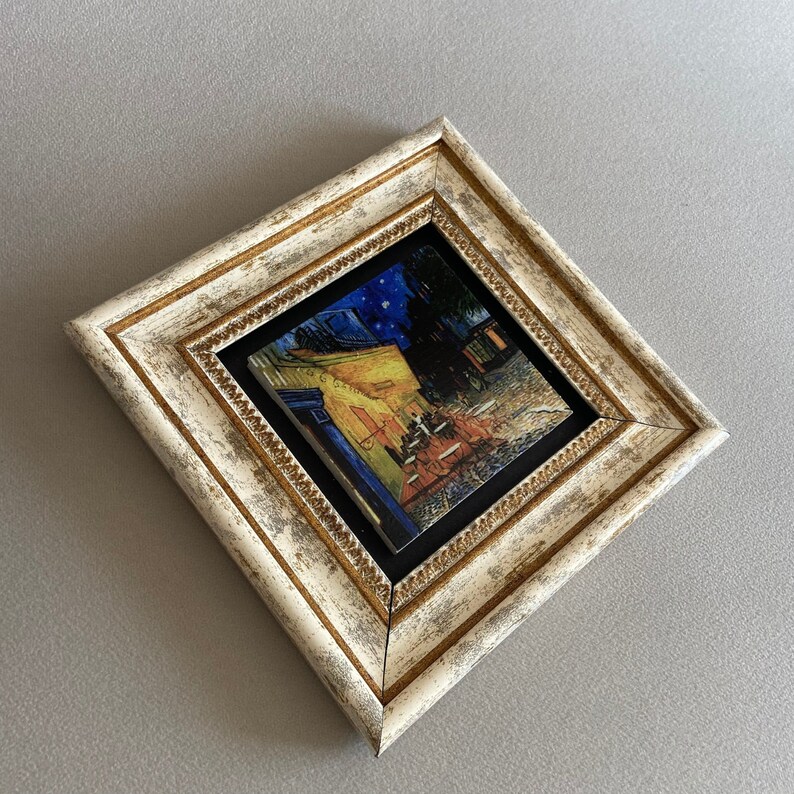 May include: A framed print of a painting depicting a cafe at night with people sitting at tables outside. The frame is white with gold accents.