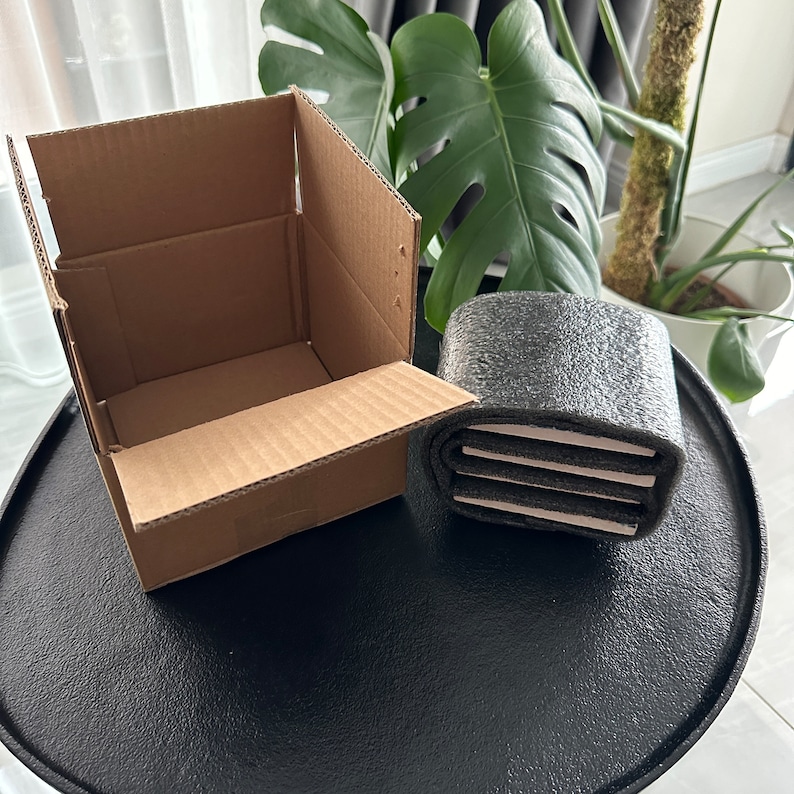 Puede incluir: Una caja de cart&oacute;n marr&oacute;n con una solapa abierta, revelando un interior vac&iacute;o. Junto a la caja hay un objeto cil&iacute;ndrico gris y blanco con una superficie texturizada.