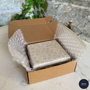 K&ouml;nnte beinhalten: Eine kleine, quadratische, graue und wei&szlig;e Keramikfliese mit geometrischem Muster ist in einem braunen Karton mit Luftpolsterfolie zum Schutz verpackt. Der Karton ist mit dem Text "BAUM ATELIER DESIGN" beschriftet.